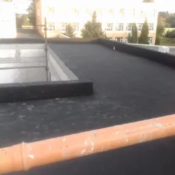 Czarna papa na płaskim dachu budynku, widoczny komin w trakcie budowy i pomarańczowa rura kanalizacyjna na pierwszym planie.