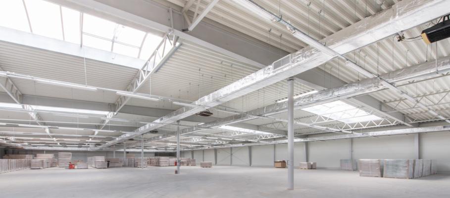 Wnętrze nowo wybudowanej hali przemysłowej z widoczną konstrukcją stalową dachu i świetlikami, przygotowanej do składowania materiałów.