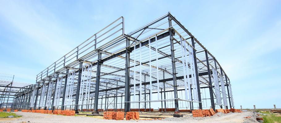 Szkielet konstrukcji stalowej hali w trakcie budowy, widoczne cegły na ziemi, błękitne niebo w tle.