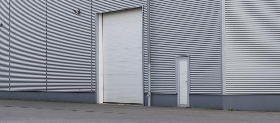 Szara hala przemysłowa z blachy falistej z bramą segmentową i drzwiami wejściowymi obok rury spustowej.