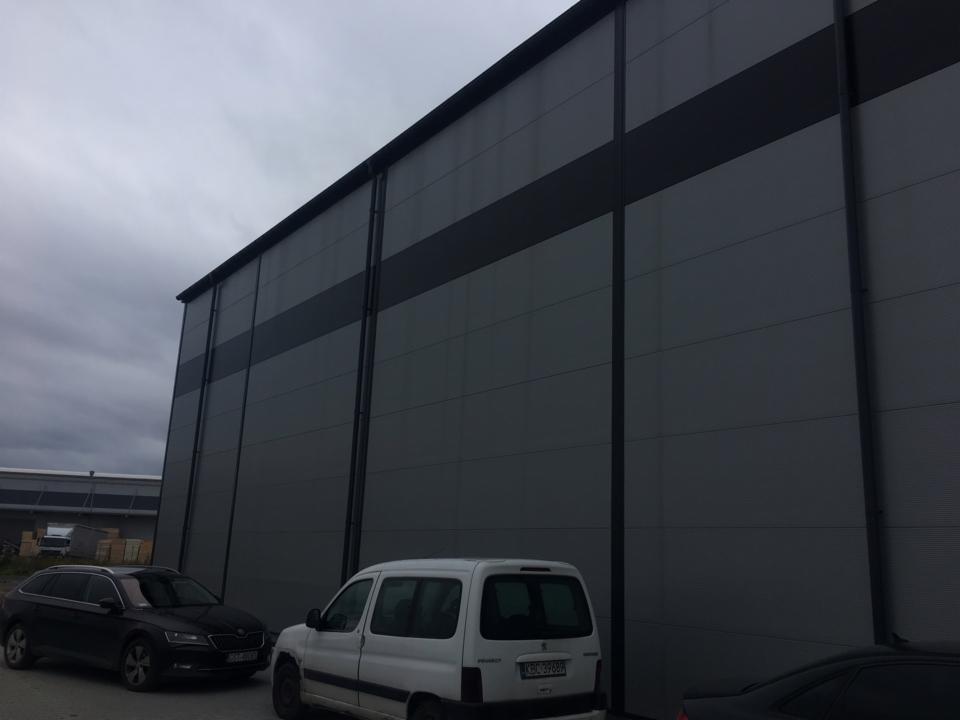 Szara hala przemysłowa z widocznymi panelami i ciemniejszym poziomym pasem, zaparkowane trzy samochody na pierwszym planie, pochmurne niebo.