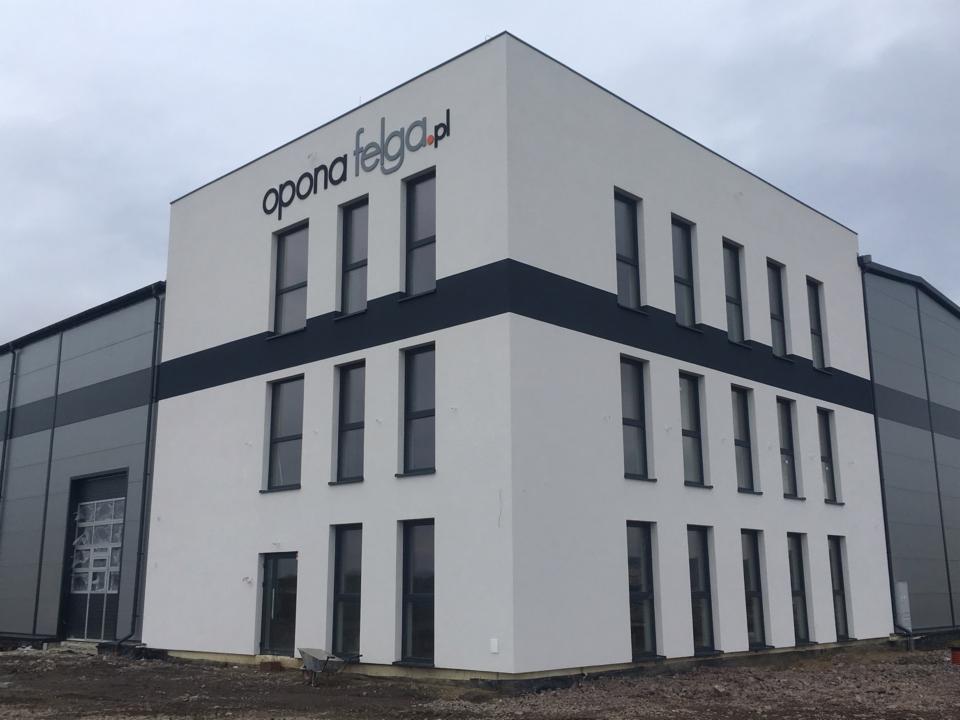 Nowoczesny budynek biurowy z przylegającą halą magazynową, świeżo wykończony, z logo firmy 'opona felga.pl' na fasadzie, widoczny teren budowy przed budynkiem.