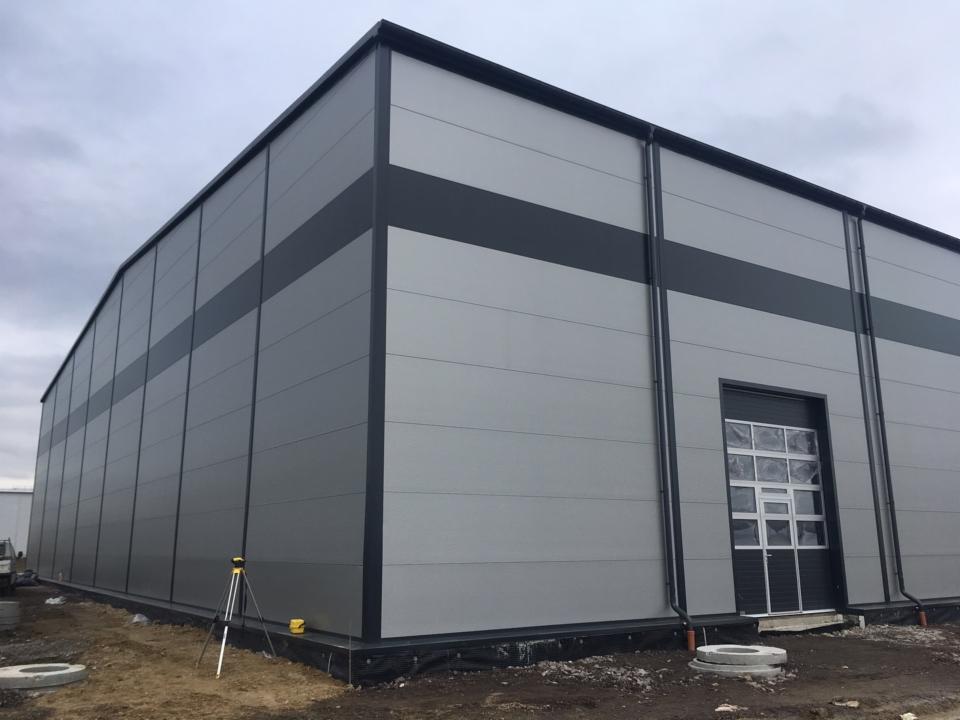 Gotowa hala przemysłowa z szarej blachy trapezowej, z automatyczną bramą segmentową z oknami, widoczny niwelator laserowy na statywie.