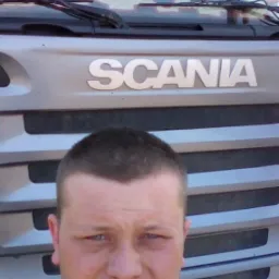 Młody mężczyzna robi selfie na tle srebrnej ciężarówki marki Scania z widocznym logo na masce i pluszowym zwierzątkiem za szybą.