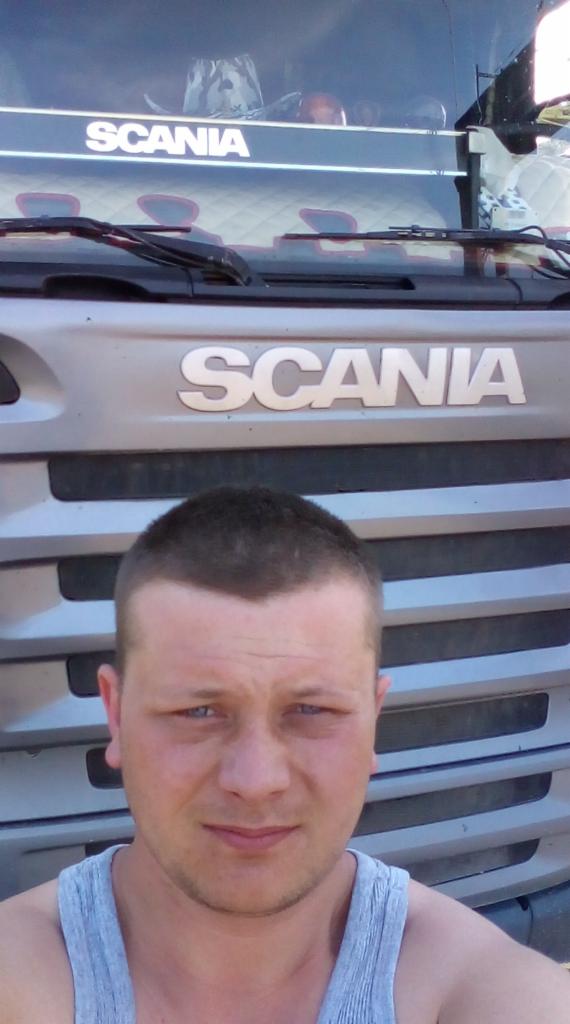 Młody mężczyzna robi selfie na tle srebrnej ciężarówki marki Scania z widocznym logo na masce i pluszowym zwierzątkiem za szybą.
