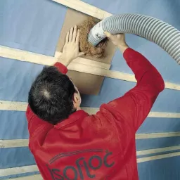 Pracownik w czerwonej bluzie z logo Isofloc aplikuje izolację celulozową do poddasza za pomocą węża, widoczne drewniane elementy konstrukcji dachu i niebieska folia paroizolacyjna.