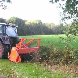Traktor z pomarańczowym mulczerem tnący krzewy przy polu w słoneczny dzień.