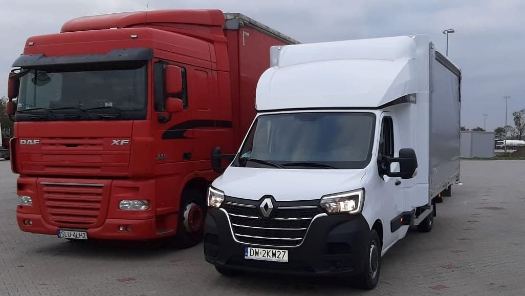 Czerwony ciągnik siodłowy DAF XF obok białego samochodu dostawczego Renault Master z zabudową kontenerową na szarym parkingu.