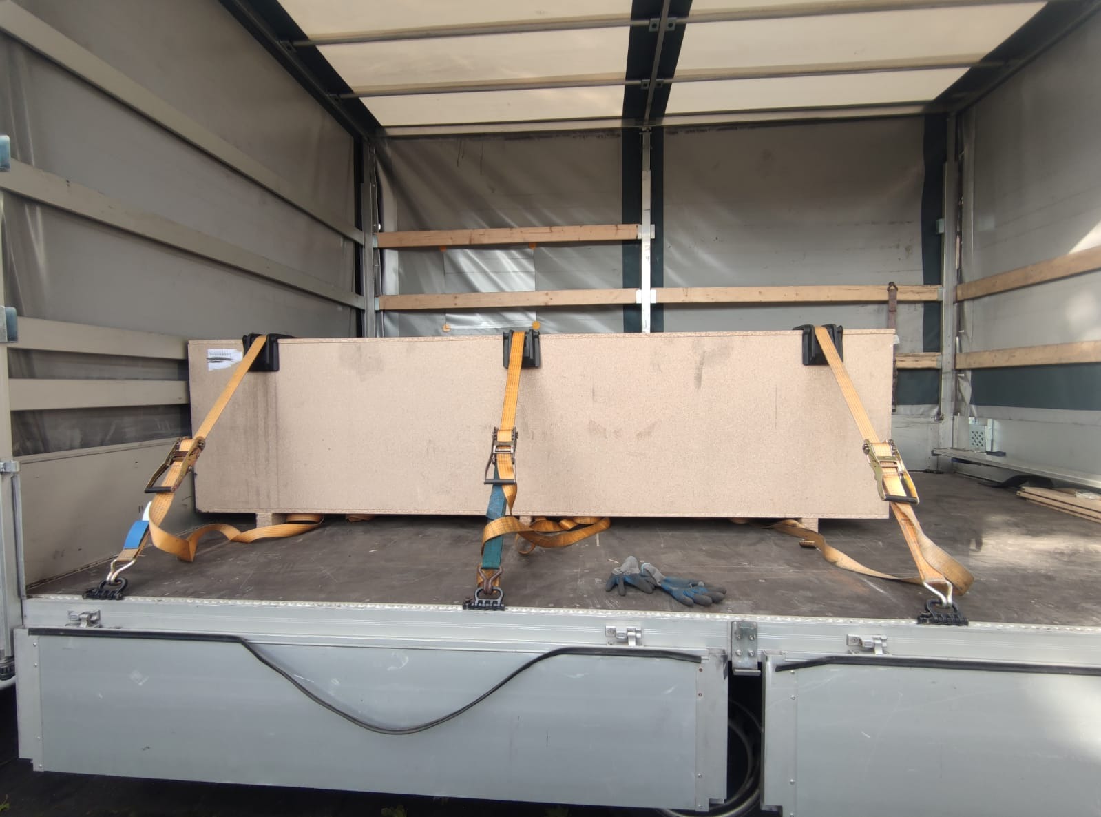 Dwie płyty meblowe zabezpieczone pasami transportowymi wewnątrz skrzyni ładunkowej samochodu ciężarowego, na podłodze leżą robocze rękawice.
