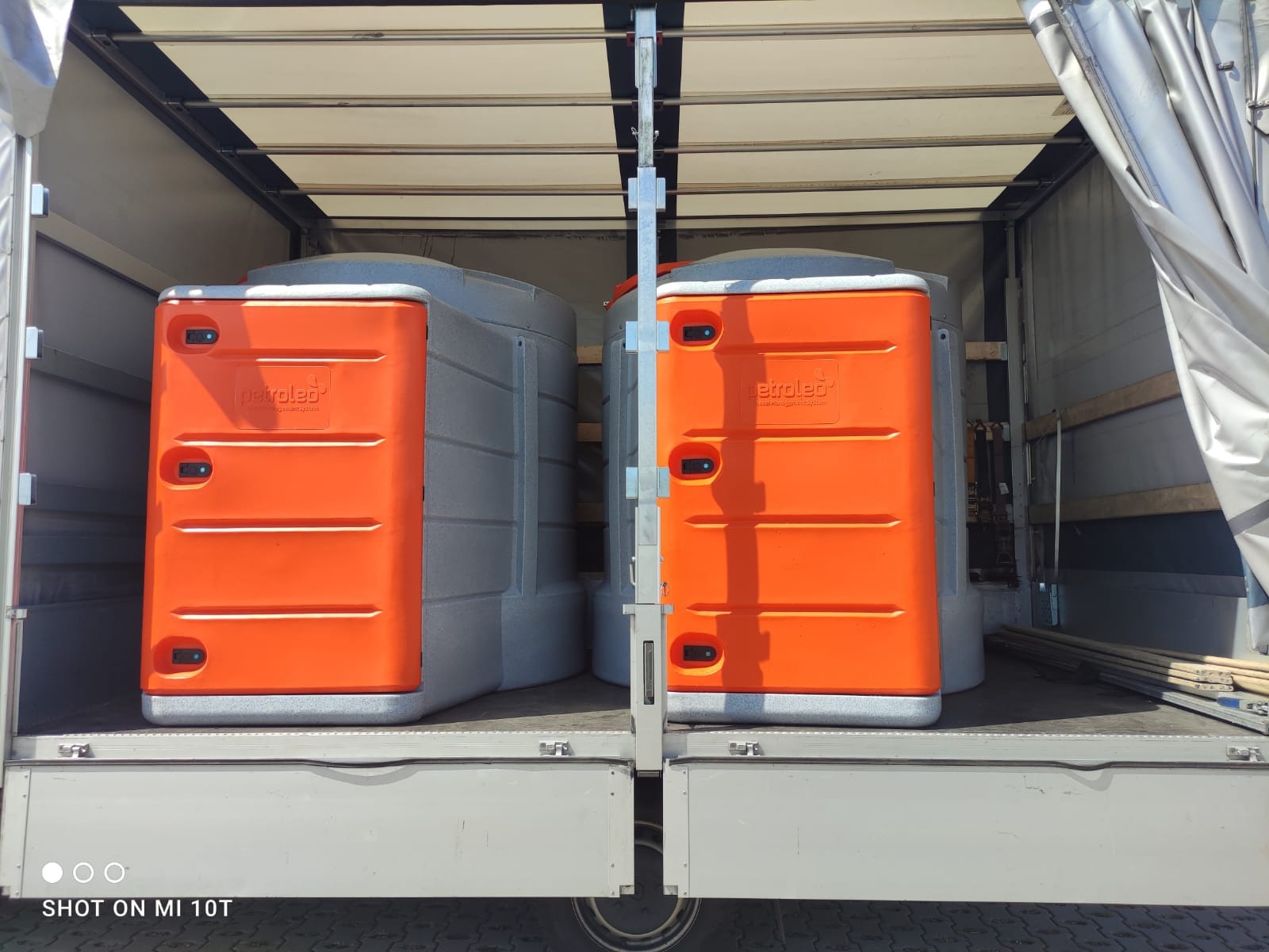 Dwie przenośne kabiny toaletowe marki Petroled, jedna pomarańczowa, druga szara, zabezpieczone wewnątrz naczepy ciężarówki podczas transportu.
