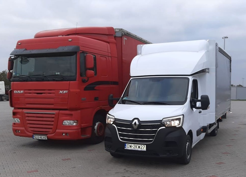 Czerwony ciągnik siodłowy DAF XF obok białego samochodu dostawczego Renault Master z zabudową kontenerową na szarym brukowanym placu.