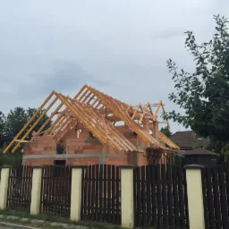 Budowa domu jednorodzinnego w trakcie: wzniesione mury z czerwonej cegły i drewniana konstrukcja dachu, widoczne zza ciemnego ogrodzenia.