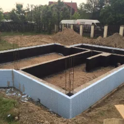 Fundament budynku w trakcie budowy, zabezpieczony czarną izolacją i obłożony styropianem, wystające pręty zbrojeniowe, w tle widoczny samochód dostawczy.