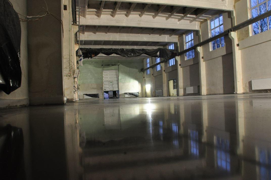 Błyszcząca, ciemnoszara posadzka w hali przemysłowej z widocznymi odbiciami okien w podłodze. W tle widoczne ściany z widocznymi instalacjami i brama wjazdowa.