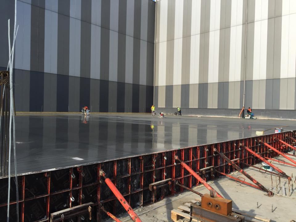 Wygładzanie świeżo wylanej, mokrej posadzki w dużej hali przemysłowej; w tle widoczna ściana hali w pionowe pasy, a w pierwszym planie deskowanie i podpory.