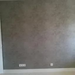 Szara ściana z efektem dekoracyjnym, biała listwa przypodłogowa, gniazdka elektryczne i uchylone białe drzwi z widoczną klamką