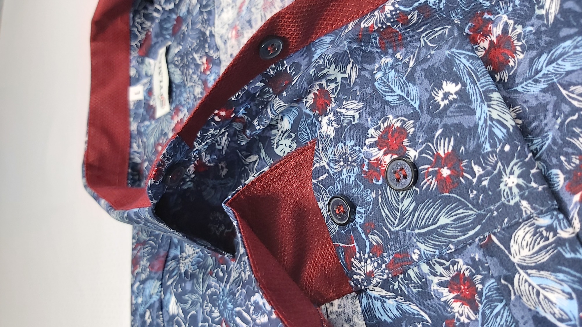Fragment męskiej koszuli z niebieskim tłem pokrytym wzorem kwiatów i liści w odcieniach bieli i czerwieni, z bordową lamówką kołnierzyka i guzikami.