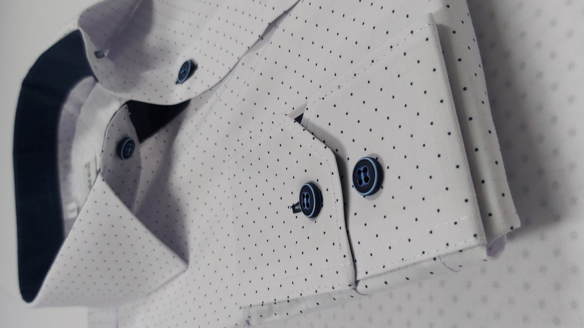 Elegancka, biała koszula męska w drobne, czarne kropki, z niebieskimi guzikami, starannie złożona, ukazująca kołnierzyk i mankiety.