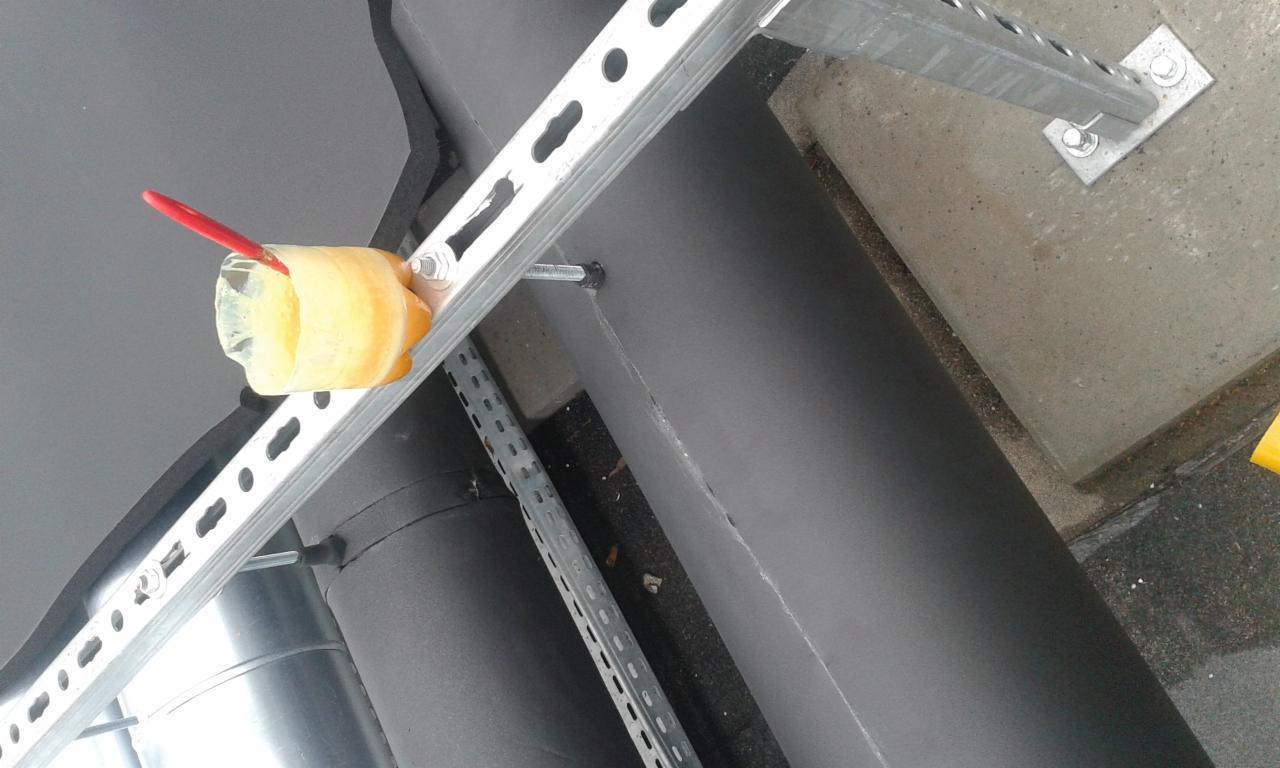 Metalowa konstrukcja wsporcza z rurami, zabezpieczona żółtą taśmą i elementem z butelki z czerwonym markerem, prawdopodobnie jako tymczasowe zabezpieczenie lub oznaczenie.