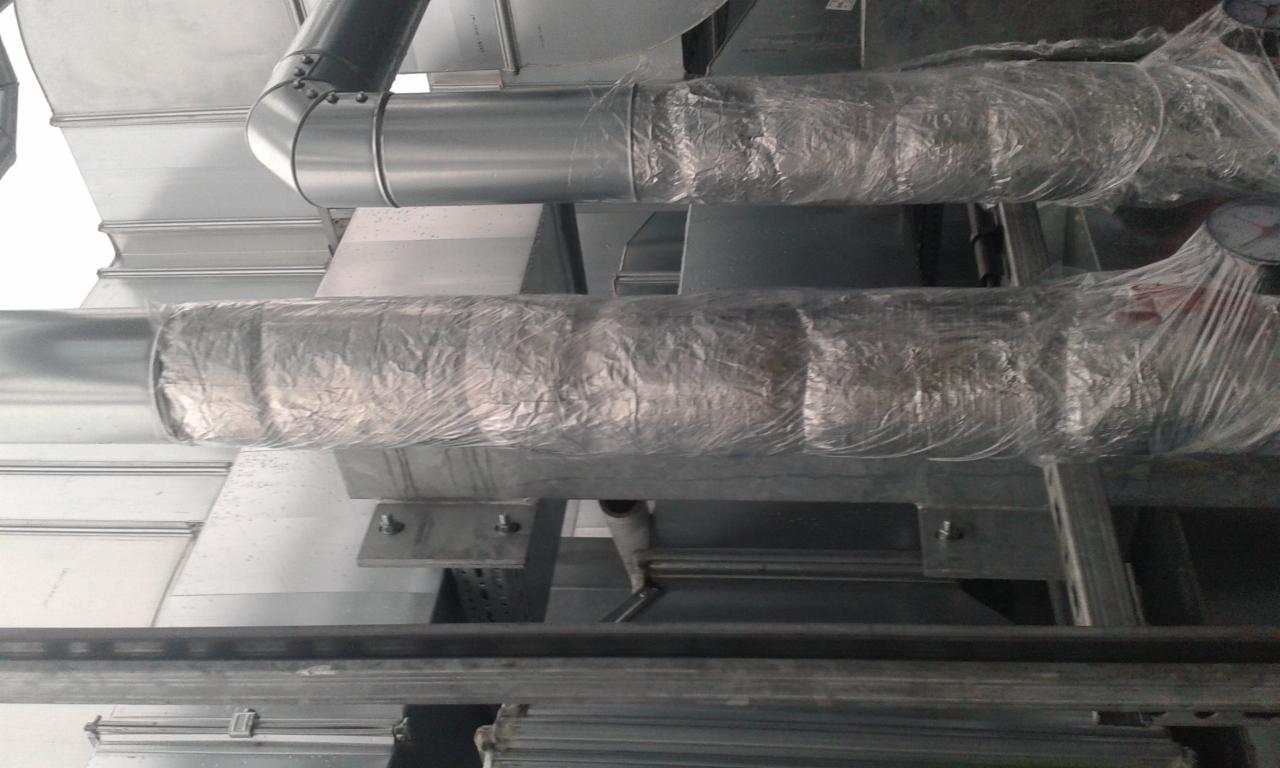 Metalowe rury wentylacyjne, częściowo owinięte folią ochronną, zamontowane pod sufitem na metalowej konstrukcji wsporczej.