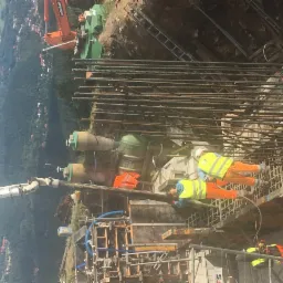 Prace zbrojarskie na budowie z widoczną konstrukcją z prętów stalowych, pracownikami w odblaskowych ubraniach oraz elementami szalunku i pompą do betonu w tle.