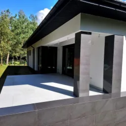 Nowoczesny taras z jasnoszarą płytką, ciemne płytki na kolumnach i ścianie, widok na dom z ogrodem w tle.