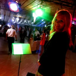 Uśmiechnięta wokalistka zespołu muzycznego z podświetloną na zielono twarzą, na tle sali bankietowej z gośćmi weselnymi i oświetleniem scenicznym.