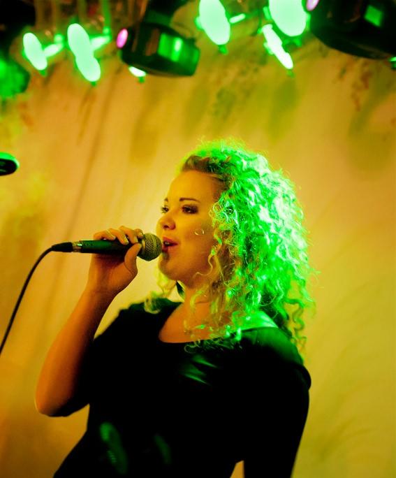 Wokalistka z kręconymi włosami śpiewa do mikrofonu na scenie oświetlonej zielonym światłem.