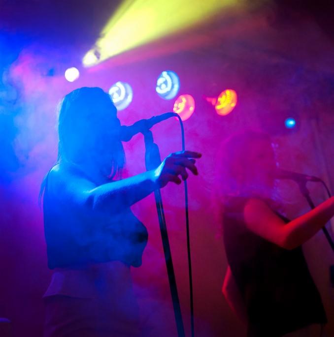 Wokalistka z mikrofonem na scenie w niebiesko-różowych światłach, drugi muzyk z tyłu, scena w lekkiej mgle.