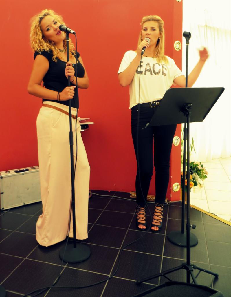 Dwie wokalistki z mikrofonami na scenie z czerwoną ścianą w tle, jedna w czarnej bluzce i szerokich, jasnych spodniach, druga w białej koszulce z napisem 'PEACE' i czarnych spodniach, obie...