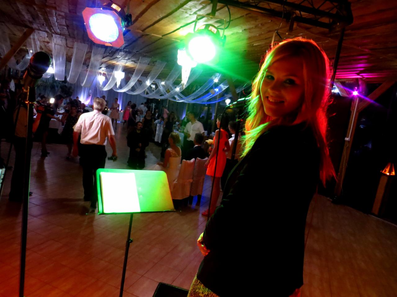 Uśmiechnięta wokalistka zespołu muzycznego z podświetloną na zielono twarzą, na tle sali bankietowej z gośćmi weselnymi i oświetleniem scenicznym.