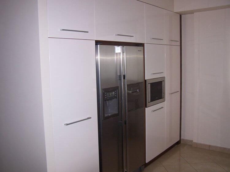 Biała zabudowa kuchenna z połyskiem, integrująca lodówkę side-by-side i mikrofalówkę, z minimalistycznymi, poziomymi uchwytami.