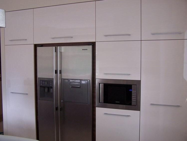 Biała zabudowa kuchenna z połyskiem, integrująca lodówkę side-by-side i mikrofalówkę, minimalistyczne uchwyty.