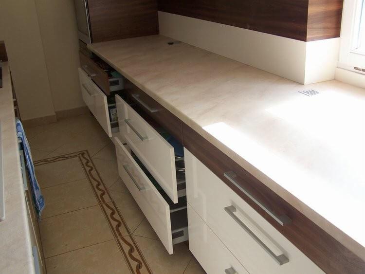 Nowoczesna zabudowa kuchenna z białymi frontami szuflad i blatem imitującym kamień, trzy szuflady częściowo wysunięte, widoczny fragment podłogi z dekoracyjnym obramowaniem.