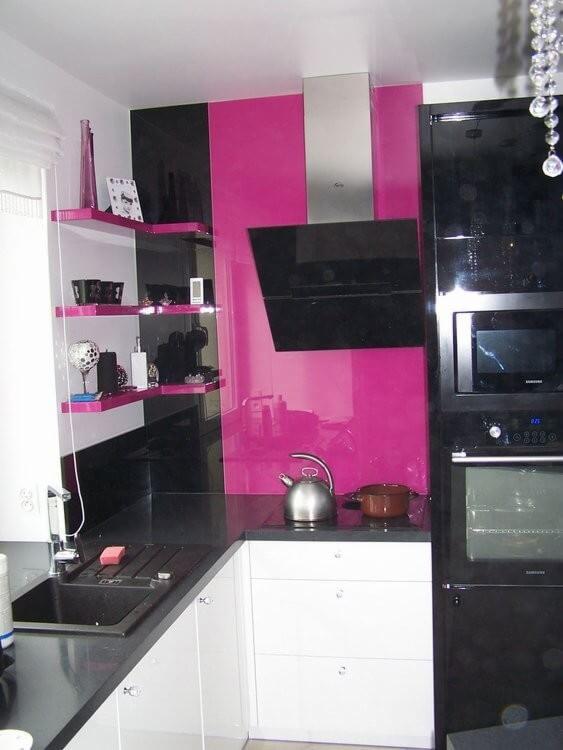 Nowoczesna kuchnia z błyszczącymi, różowymi panelami ściennymi, czarnymi szafkami i srebrnym okapem, uzupełniona białymi szafkami dolnymi i czarnym blatem.