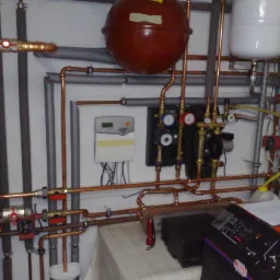 Zawiła instalacja grzewcza z miedzianych rur, zbiornikami wyrównawczymi i pompami obiegowymi, widoczna na tle białej ściany z szarymi rurami. Na pierwszym planie fragment stołu z dokumentacją...