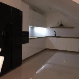 Nowoczesna kuchnia w skosach, białe szafki z drewnianym blatem, czarny zlew i bateria, czarna lodówka side-by-side, płytki podłogowe w odcieniach beżu.