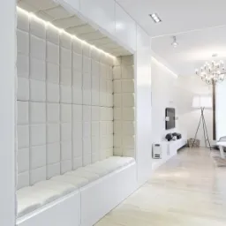 Biała, tapicerowana wnęka z siedziskiem, wbudowana w ścianę, z oświetleniem LED u g&oacute;ry, widok na salon z żyrandolem.