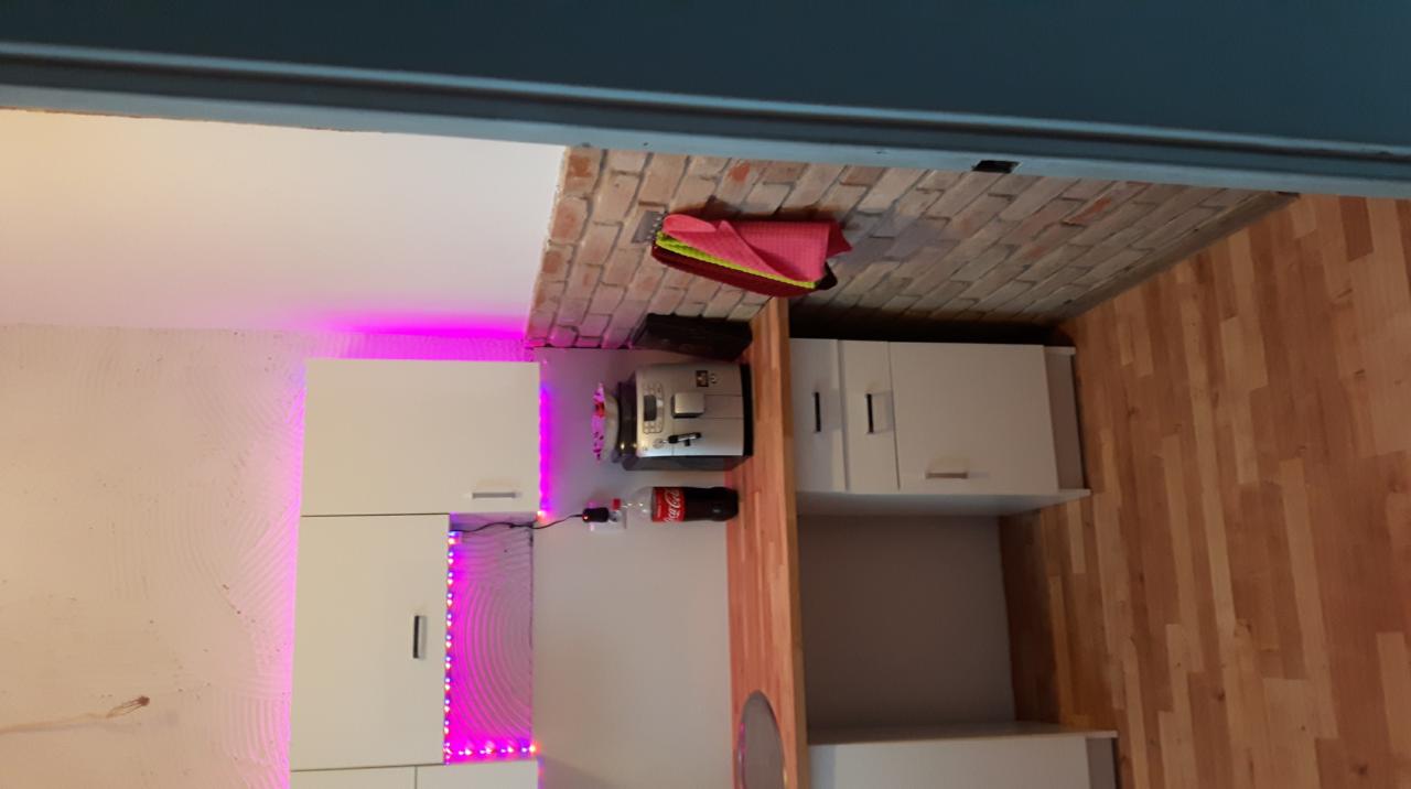 Nowoczesna kuchnia z białymi szafkami, oświetleniem LED w kolorze fioletowym, blatem imitującym drewno, ekspressem do kawy, butelką Coca-Coli i ceglaną ścianą w tle.