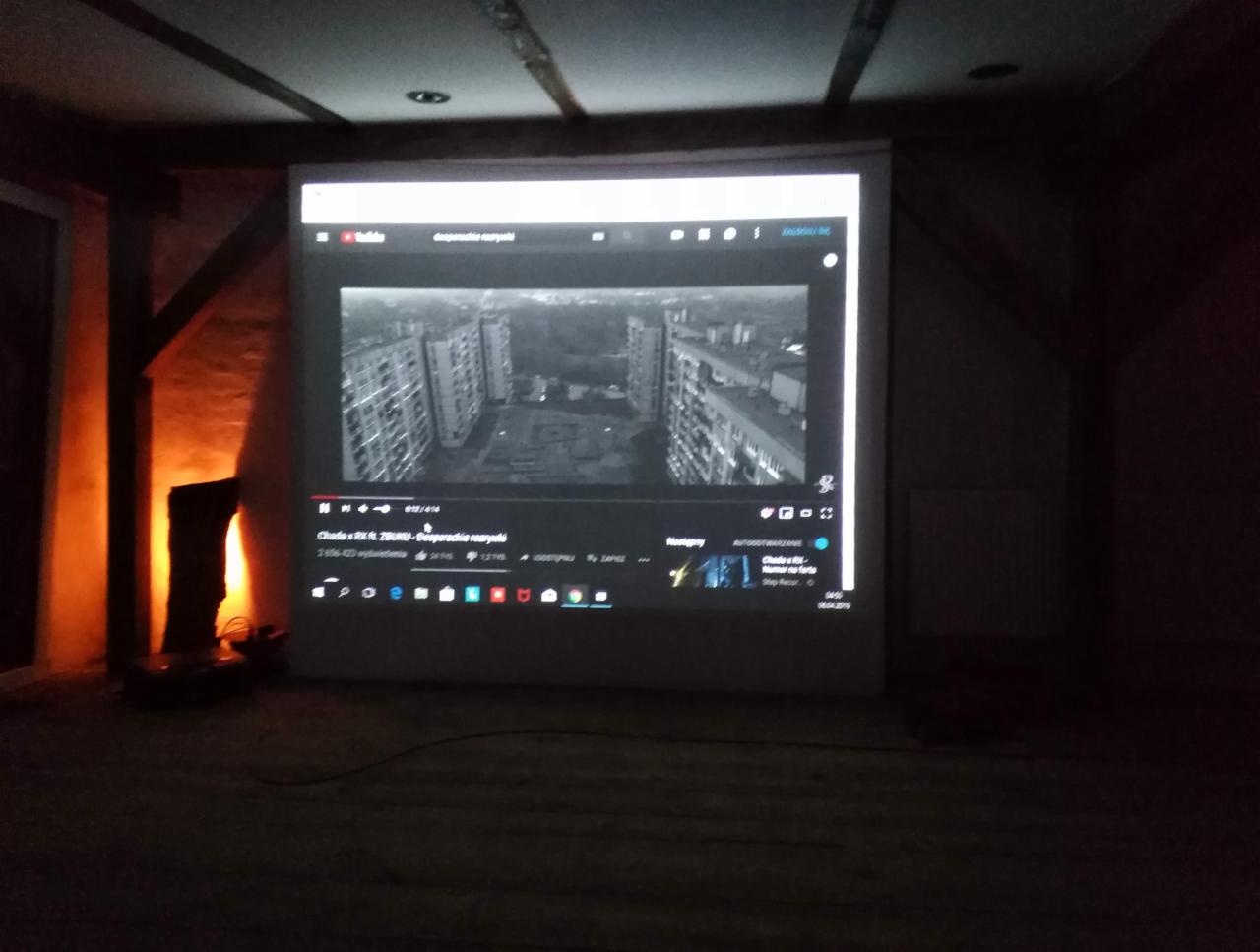 Projekcja filmu na ścianie w pokoju z drewnianym stropem i pomarańczowym oświetleniem, widoczny interfejs YouTube z filmem o blokach.