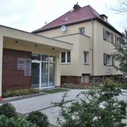 Dwukondygnacyjny budynek murowany z kremową elewacją, brązowymi okiennicami i czerwonym dachem, z dobudowanym parterowym wejściem z drewnianą okładziną i szklanymi drzwiami, otoczony brukowanym...