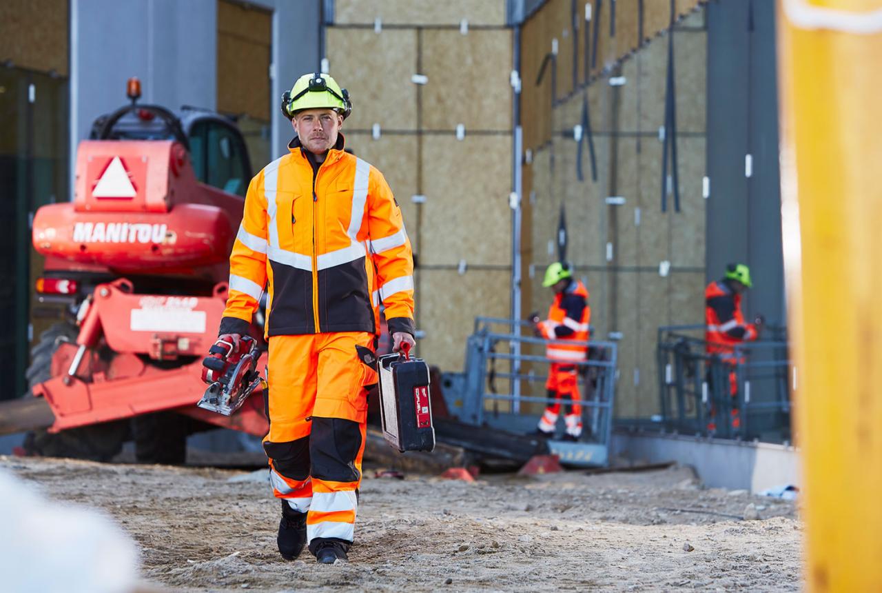 Pracownik budowlany w odblaskowym ubraniu, kasku z lampką czołową, niosący skrzynkę narzędziową i piłę tarczową, na tle budowy z podnośnikiem i maszyną Manitou.