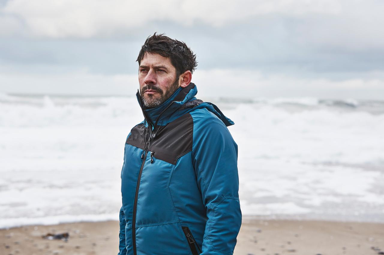 Mężczyzna w niebieskiej kurtce na tle wzburzonego morza, spoglądający w dal.