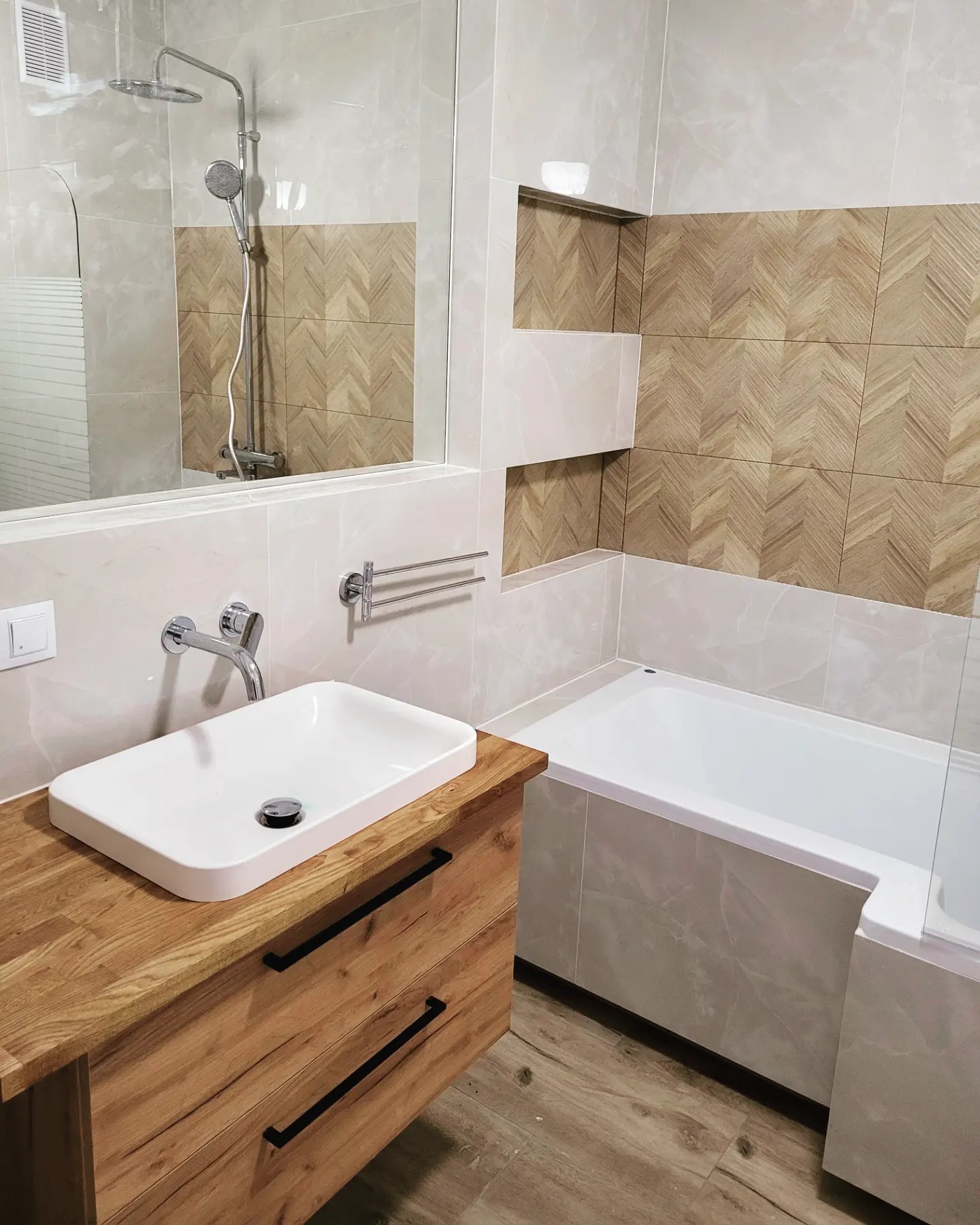 Nowoczesna łazienka z drewnianą szafką pod umywalką, płytkami imitującymi drewno na ścianie i wanną obudowaną płytkami w odcieniach szarości.