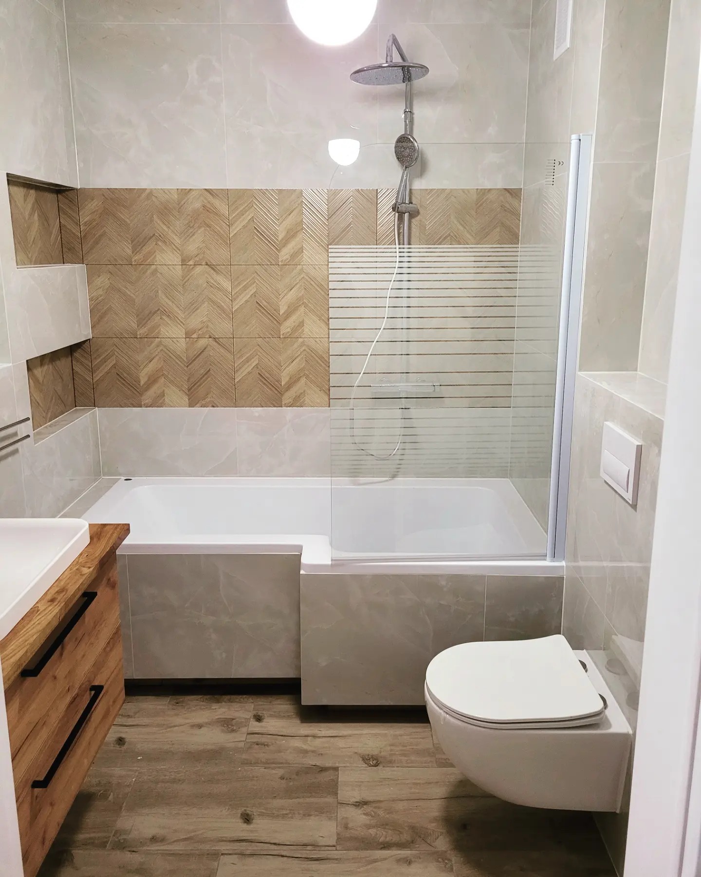 Nowoczesna łazienka z wanną obudowaną płytkami imitującymi marmur, drewnopodobnymi płytkami ułożonymi w jodełkę na ścianie oraz podwieszaną toaletą.