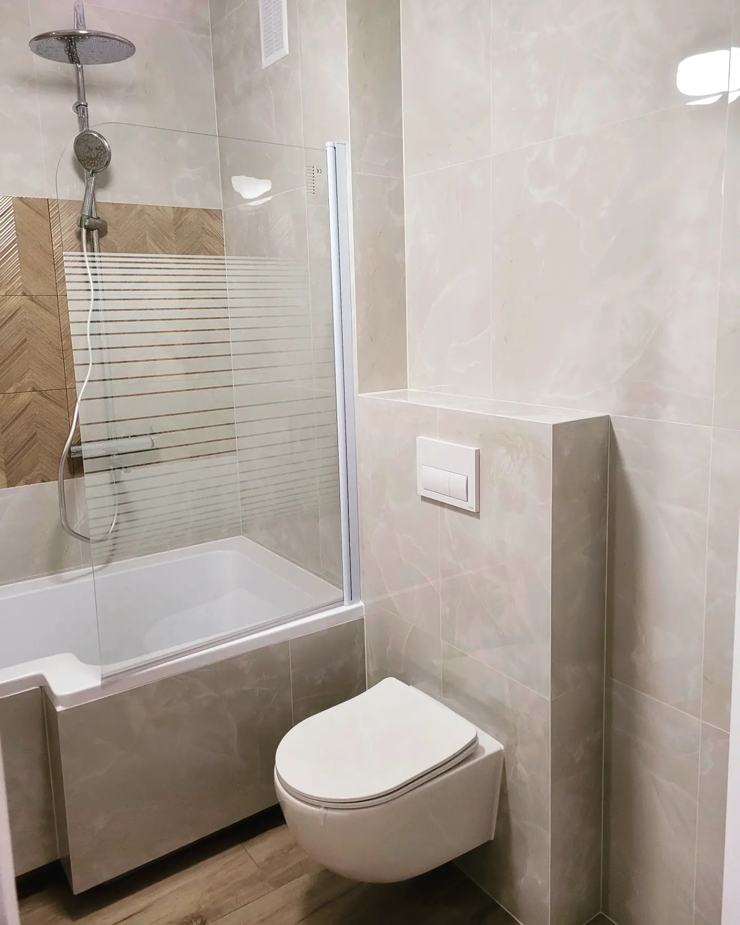 Wykończona łazienka z beżowymi płytkami imitującymi marmur, kabiną prysznicową z poziomymi paskami i wiszącą toaletą.