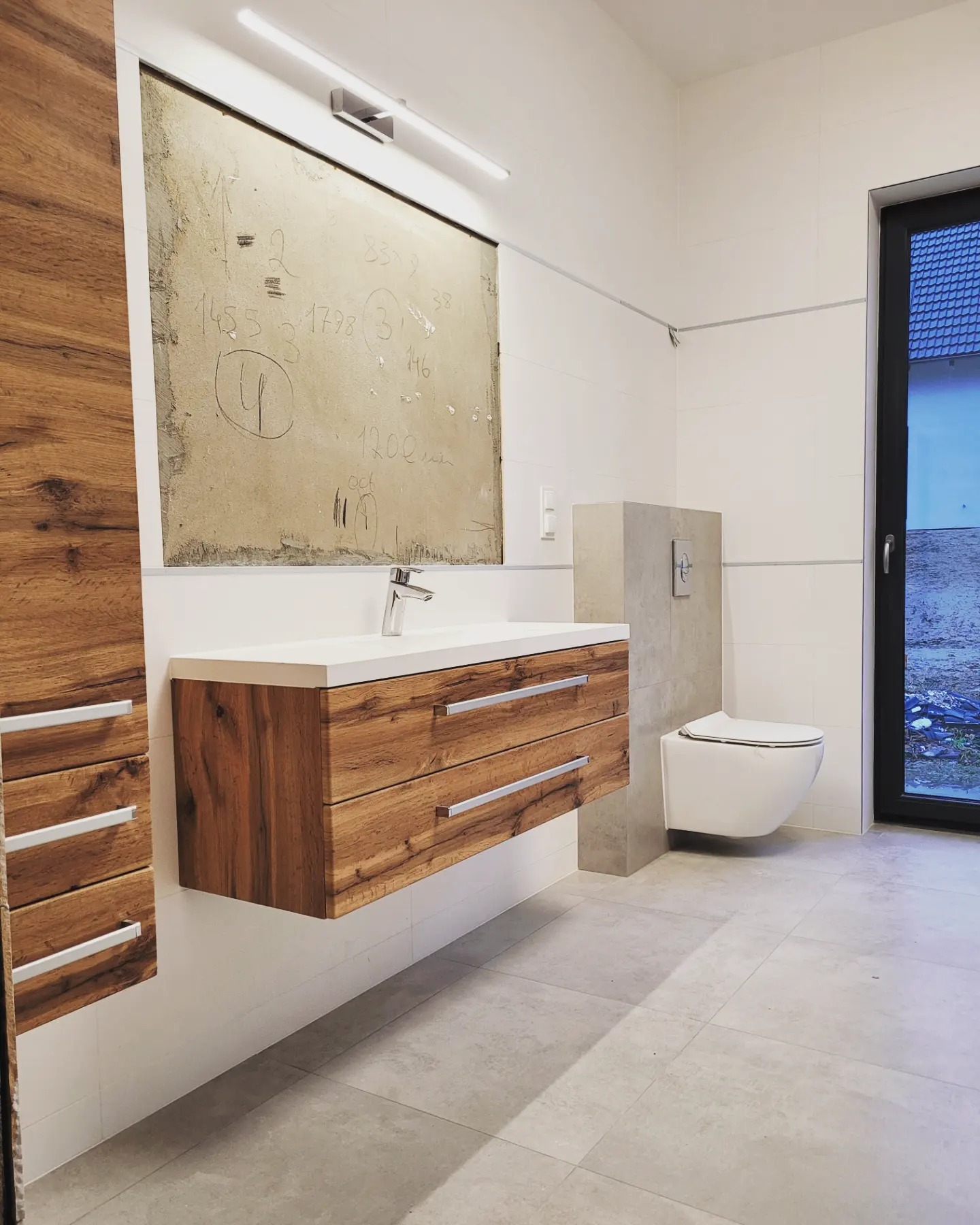 Wnętrze łazienki w trakcie prac: wisząca szafka pod umywalkę z drewnianymi frontami, biała umywalka, wisząca toaleta, ściany z białych płytek, fragment ściany przygotowany pod lustro z widocznymi...