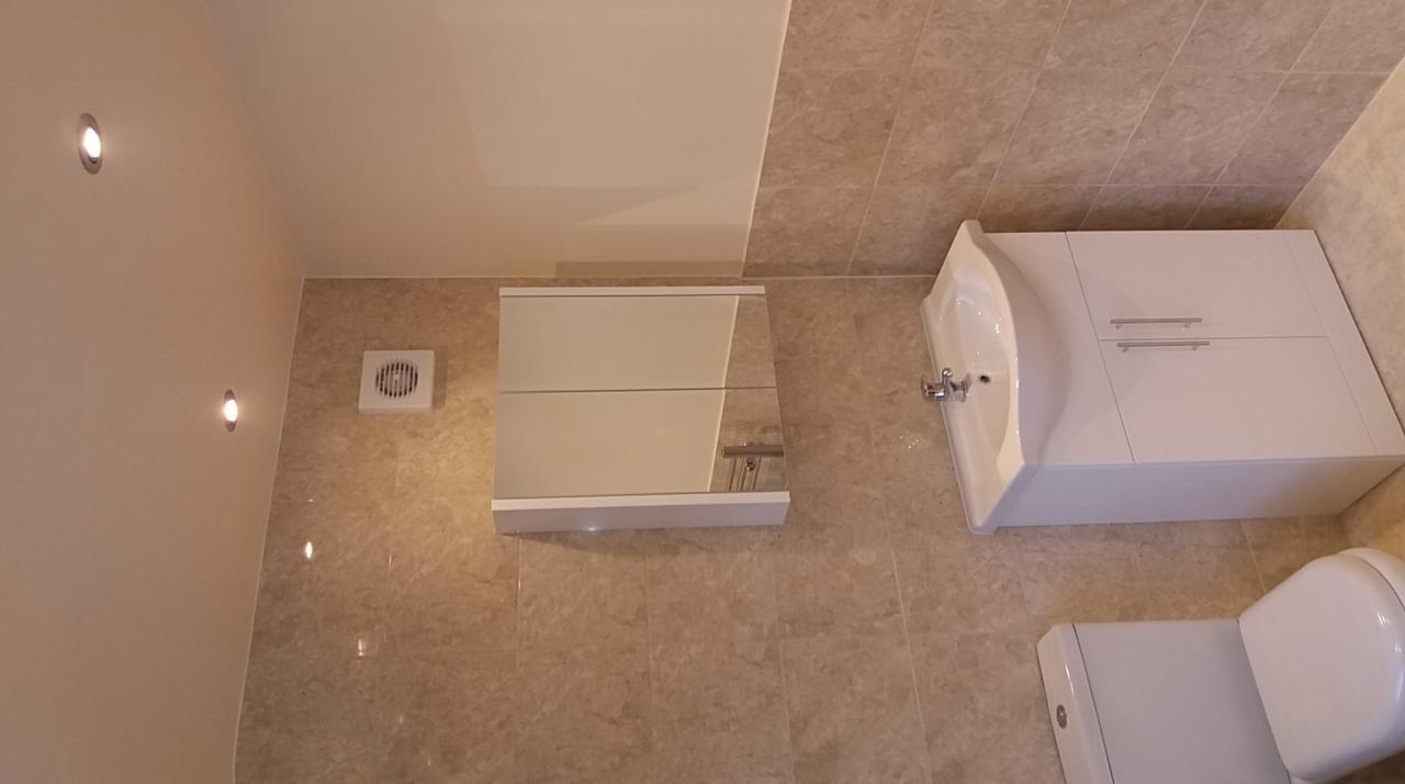 Wykończona łazienka z beżowymi płytkami, białą umywalką z szafką, toaletą i lustrzaną szafką na ścianie, oświetlona punktowymi lampami sufitowymi.