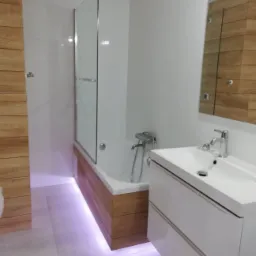 Nowoczesna łazienka z podświetleniem LED pod wanną obudowaną drewnopodobnymi panelami, białą umywalką z szafką i lustrem w drewnianej ramie.