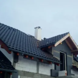 Częściowo pokryty ciemną dachówką dach domu w trakcie budowy, widoczny komin, część dachu pokryta śniegiem, elementy drewniane konstrukcji dachowej.